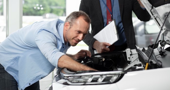 Two mechanics in a garage discuss while one holds a clipboard, referencing the California Lemon Law, and the other a wrench. A car with an open hood, potentially falling under used cars regulations, stands in the background.
