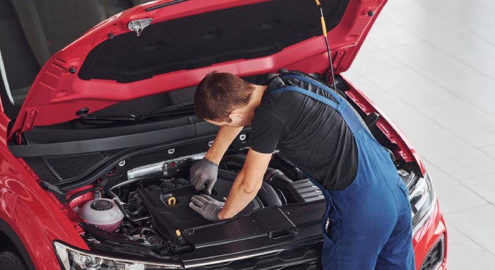 Two mechanics in overalls discuss near a silver car; one holds a clipboard. In the background, another car with an open hood awaits attention, possibly dealing with CA lemon law issues.