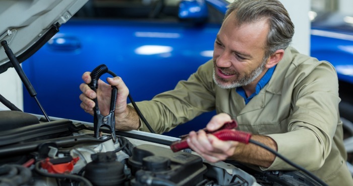 A mechanic in a beige jacket uses jumper cables to revive a car inside the garage, reminiscent of the precision and determination seen in a Lemon Law attorney's pursuit of justice. In the background, a blue vehicle stands as if waiting its turn for courtroom success.