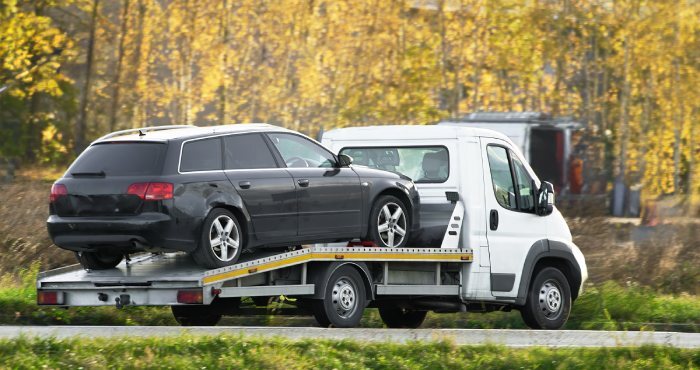 A black station wagon, possibly subjected to California Lemon Law issues, is being transported on a flatbed truck. The scene unfolds in a rural area, with trees and grass painting the backdrop.