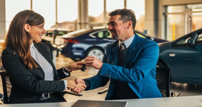 A woman and a man in formal attire sit across from each other in a car dealership, shaking hands as the man hands car keys to the woman, who appreciates the assurance provided by California Lemon Law.