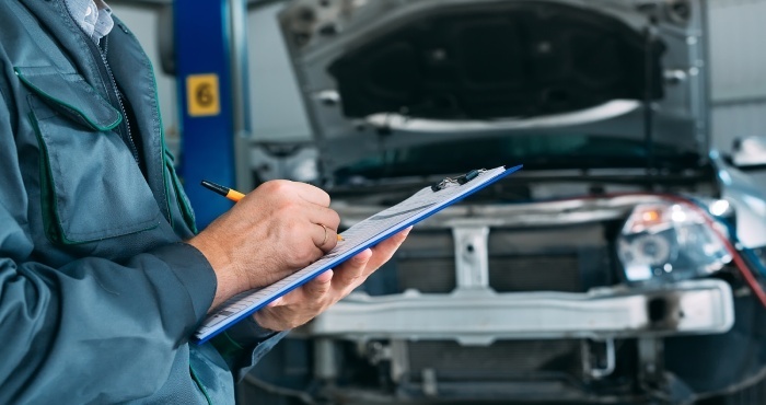 Mechanic holding a clipboard, inspecting a car with an open hood in a garage, ensuring everything is up to par to avoid any future lemon law attorney visits.