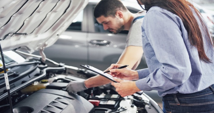 A mechanic examines a car engine while a woman, holding a clipboard, makes notes about potential vehicle modifications.