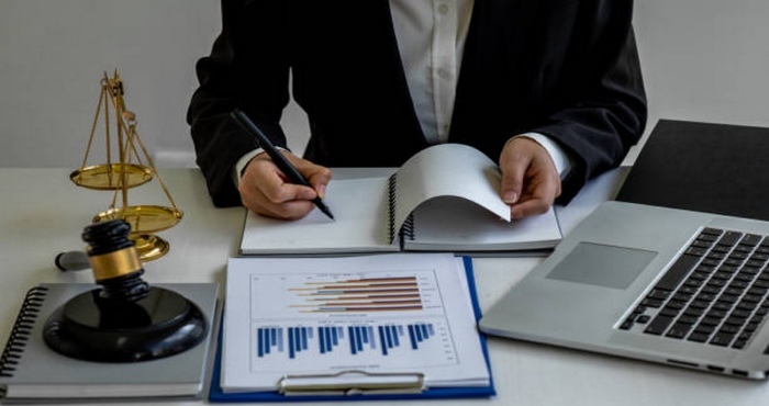 A person in a suit diligently writes in a notebook at a desk adorned with a laptop, legal scales, and charts. This vibrant scene resonates with the dedication of lemon law attorneys working towards courtroom success.