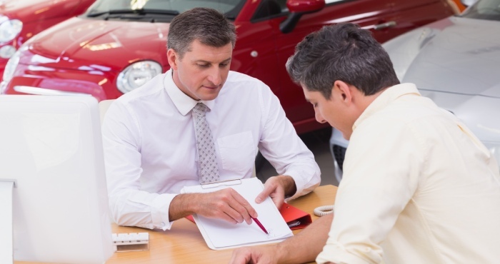 Two men sit at a desk in a bustling car dealership. One man, resembling a seasoned Lemon Law attorney, gestures confidently with a pen towards the document as the other listens attentively, perhaps pondering courtroom success. Red cars gleam enticingly in the background.