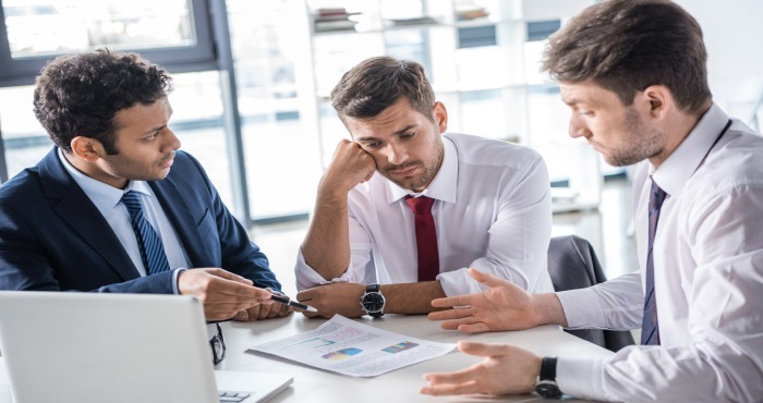 Three men in business attire discuss documents with charts at a table, clearly analyzing case results. One looks frustrated while the others gesture and talk, perhaps strategizing for future courtroom success. A laptop is open in the foreground, displaying critical data.