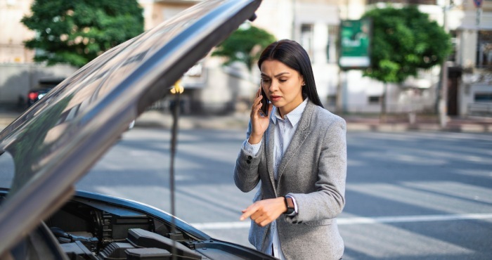 A woman in a gray blazer uses her smartphone to inspect the engine of a car with the hood open on a bustling California city street, mindful of potential Lemon Law implications.