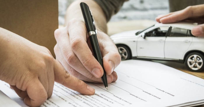 Hands sign a document with a pen, as a miniature white car sits on the table, highlighting the California Lemon Law's impact.