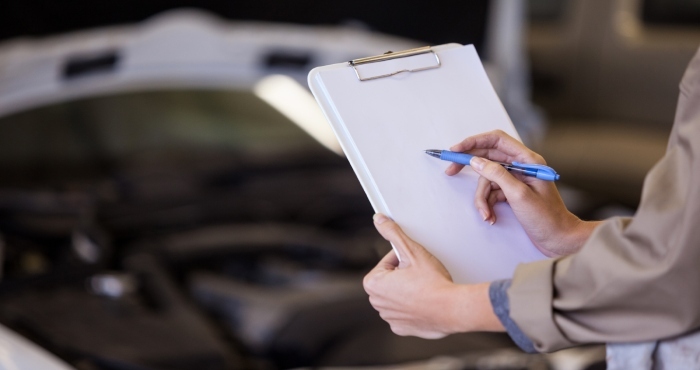 A person holding a clipboard and pen stands in front of a car with an open hood, likely assessing whether it qualifies under California's Lemon Law.