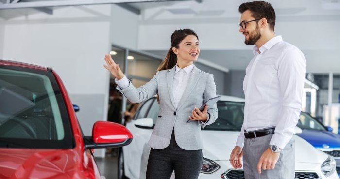 A woman in a gray blazer chats with a man in a white shirt beside a red car at a California showroom, possibly discussing consignment sales details.