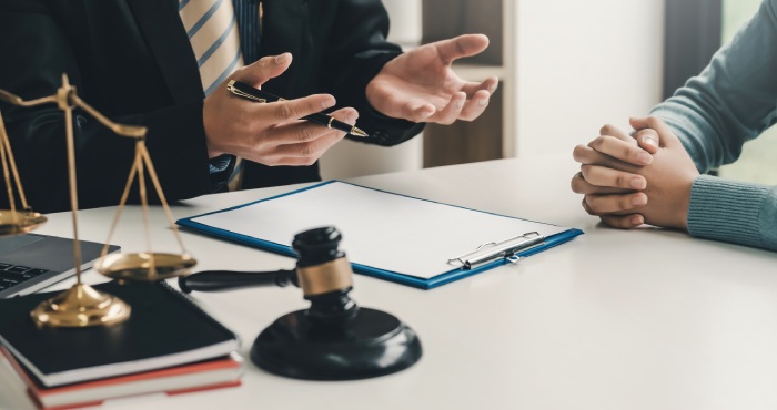 Two people are seated at a table with legal documents, a gavel, and scales. One person gestures with a pen, discussing the recent overhaul of the California Lemon Law, while the other listens intently, hands clasped.